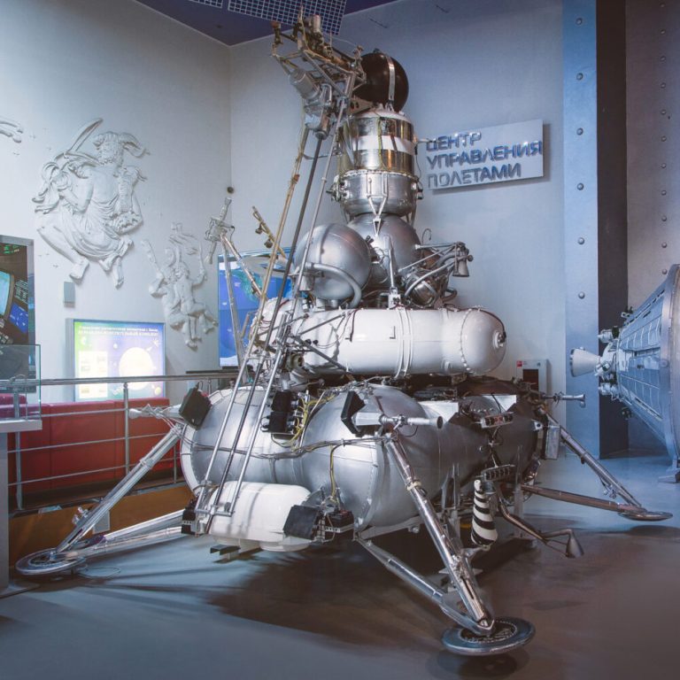 A large, complex silver spacecraft composed of stacked spherical tanks, cylindrical parts, and extended landing legs stands in the center of an indoor display room. The scene is brightly lit, highlighting the metallic sheen of the structure against smooth grey flooring and light blue-grey walls. White relief sculptures and a sign with Cyrillic text appear on the wall in the background to the left and rear.