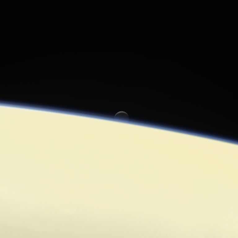 A pale yellow, smoothly textured sphere fills the bottom half of the frame, slanting downward to the right. A thin, vivid blue outline traces its curved edge against a solid black space background. A tiny, dim grey crescent moon peeks out from directly behind the blue horizon near the center.