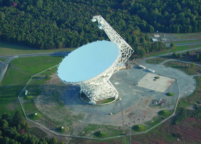 An aerial view shows a massive, white dish structure tilted upward in the center, supported by a dense lattice of metal scaffolding. A tall white support arm extends high above the dish toward the top frame. The structure sits on a wide, cleared grey clearing surrounded by green grassy areas and a thick border of dark green forest.