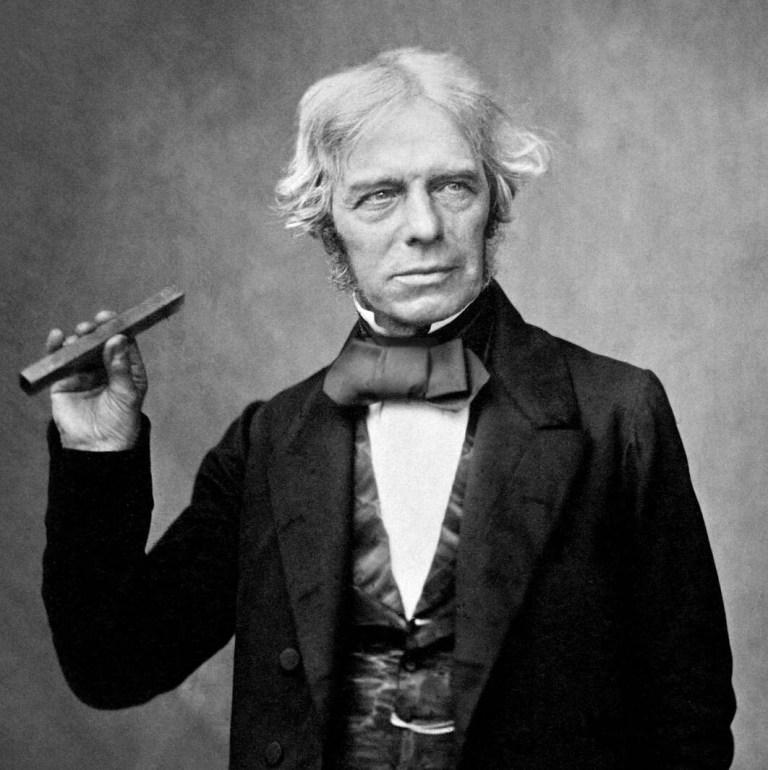 A black-and-white portrait shows a man with wavy, light-colored hair and sideburns standing in three-quarter view against a plain grey studio background. He wears a dark suit jacket, a vest, a white collared shirt, and a wide black bow tie, holding a small rectangular bar horizontally between his fingertips in his raised right hand. Soft key lighting highlights his facial features and shirt front, creating moderate contrast with his dark clothing.