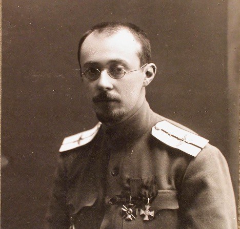 A sepia-toned bust portrait features a man with wire-rimmed spectacles, thin hair, a mustache, and a goatee, looking directly forward. He wears a high-collared military tunic adorned with light-colored epaulets on both shoulders and small medals pinned to his left chest pocket. The subject is evenly illuminated against a smooth, dark, monochromatic backdrop.