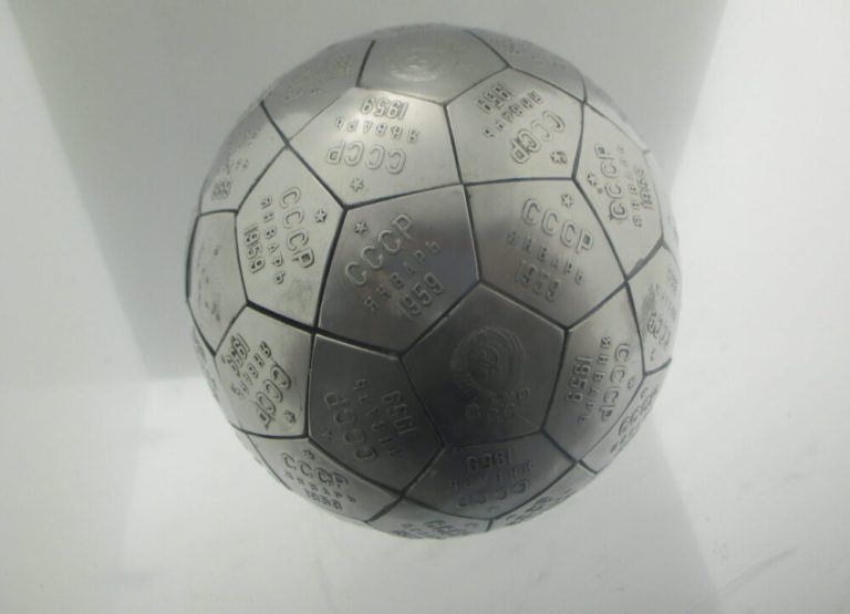 A metallic grey sphere made of interlocking pentagonal and hexagonal tiles sits centrally against a bright, plain white background. Each brushed-metal tile is engraved with Cyrillic text, numerals, and small crest emblems, resembling a soccer ball structure. Overhead display lighting creates soft reflections on the upper hemisphere and subtle shadows along the lower curves.