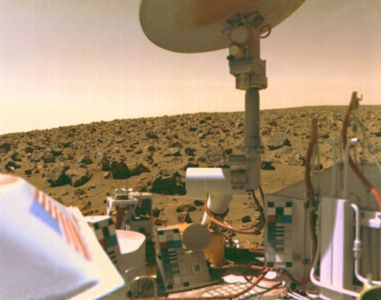 A close-up view of mechanical equipment and a white dish antenna fills the right side and foreground of the frame, bathed in warm, yellowish-orange light. In the background, a vast, flat landscape covered in small, dusty rocks stretches toward a low horizon under a hazy, pale sky. Light contrast and muted earth tones dominate the scene, emphasizing the dry, rocky terrain.