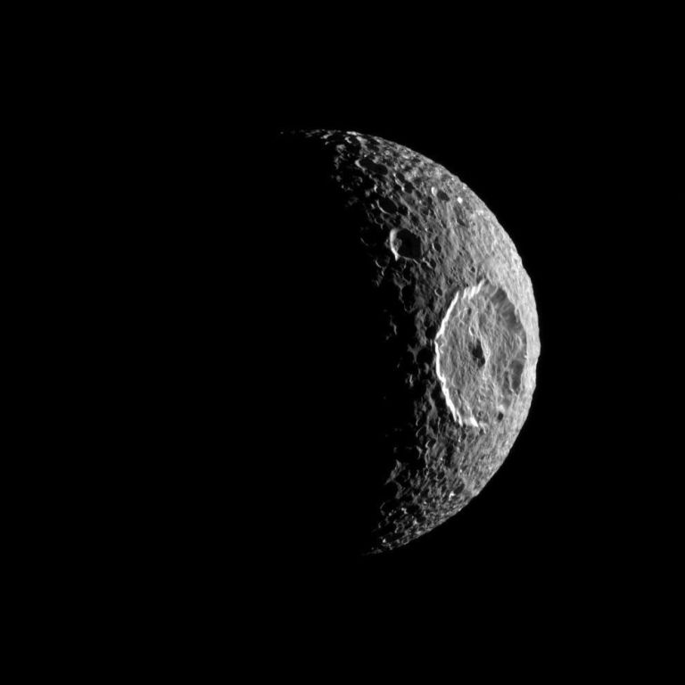A black-and-white photograph shows a crescent moon-like body positioned on the right side of the frame against a solid black space background. Strong side lighting illuminates its heavily cratered grey surface, featuring a prominent, large circular crater with a bright raised rim near the center edge. The left half of the spherical body vanishes completely into deep shadow, creating high visual contrast.
