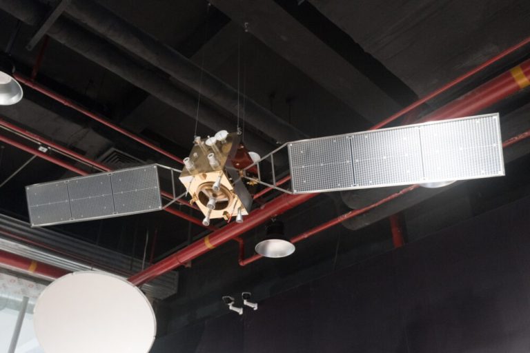 A metallic, box-shaped satellite model hangs suspended in the center against a dark industrial ceiling crossed with red pipes. Two large, grid-patterned rectangular solar panels extend outward from opposite sides like wings, while small circular dishes and thruster nozzles attach to its brassy body. Direct overhead lighting reflects off the metallic surfaces and the white, grid-textured solar panels, creating moderate contrast with the shadowy ceiling behind it.