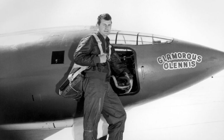 A black-and-white photograph shows a pilot standing proudly next to the nose of an airplane, holding a helmet in his left hand and a parachute pack slung over his shoulder. The text "GLAMOROUS GLENNIS" is painted in large letters on the aircraft's curved nose just ahead of the open cockpit door. Bright daylight illuminates the scene, creating a sharp contrast between the dark flight suit and the light background.