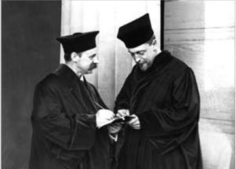 A black-and-white photograph shows two men standing side by side in dark, flowing academic or ceremonial robes and matching, structured black caps. The man on the left, who has a mustache, holds a small notebook while writing in it with a pen, and both men look down at the pages together. They stand against a light-colored wall that creates high contrast with their dark attire.