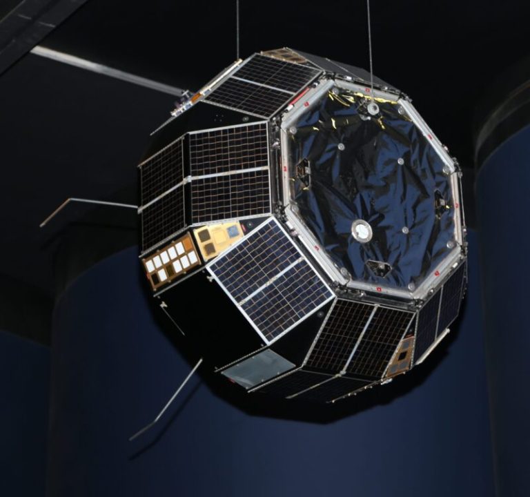 A multi-sided, pumpkin-shaped satellite hangs suspended in the center of the frame against a dark indoor background. Its dark exterior features faceted black solar panels bordered by silver frames, small gold-colored detail panels, and several thin metal antennas extending outward from its sides. High overhead lighting reflects brightly off the metallic trim and the crinkled, dark foil-like material covering its circular top surface.