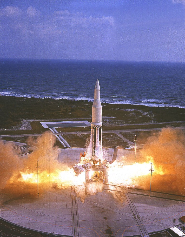 A tall, vertical white rocket lifts off from a concrete launchpad, surrounded by massive plumes of bright orange and white exhaust smoke. The launchpad sits near a coastline with dark green vegetation and blue ocean waves stretching across the horizon beneath a pale blue sky. Bright light and high contrast illuminate the base of the rocket and the expanding smoke cloud against the surrounding landscape.