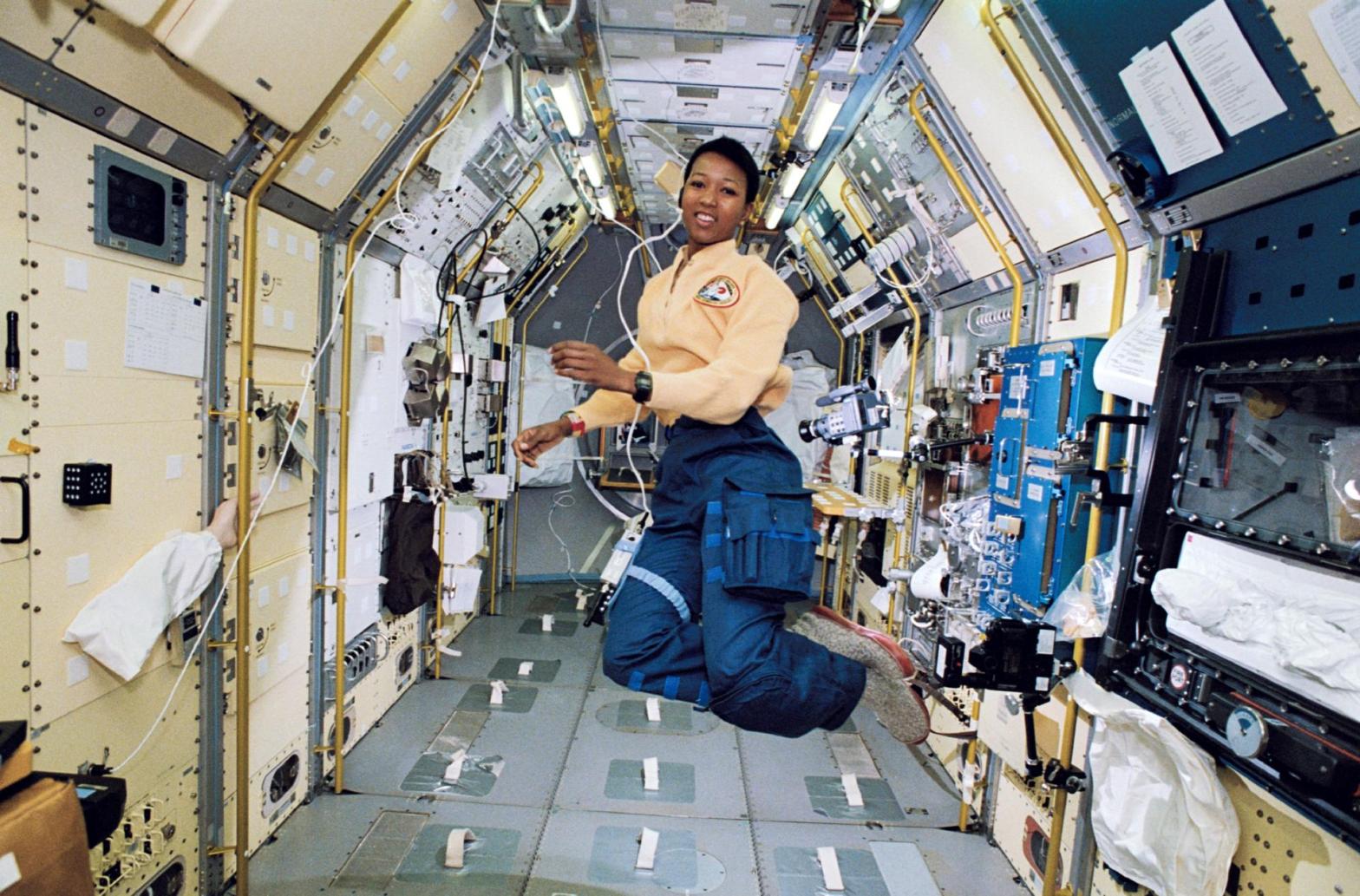 A woman in a yellow top and blue trousers floats mid-air in the center of a narrow, tunnel-like spacecraft corridor lined with modular white panels, equipment racks, and yellow handrails. Bright, even interior overhead lights illuminate her and the surrounding laboratory modules, creating clear visibility throughout the enclosed space. She smiles toward the camera as she hovers freely above the metallic floor grid.
