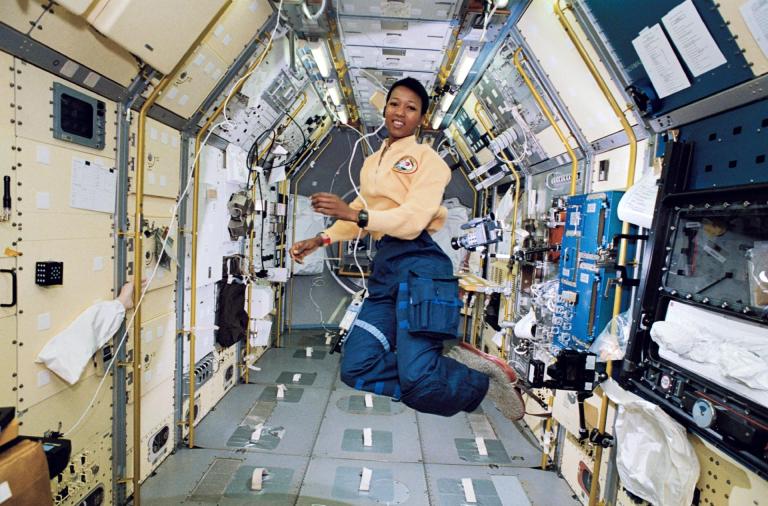 A woman in a yellow top and blue trousers floats mid-air in the center of a narrow, tunnel-like spacecraft corridor lined with modular white panels, equipment racks, and yellow handrails. Bright, even interior overhead lights illuminate her and the surrounding laboratory modules, creating clear visibility throughout the enclosed space. She smiles toward the camera as she hovers freely above the metallic floor grid.
