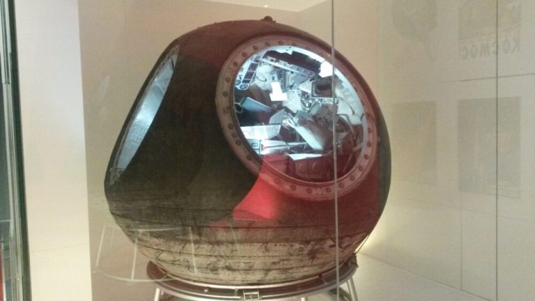 A large, spherical space capsule rests on a low ring stand inside a glass display case against plain white museum walls. The capsule’s charred exterior features patches of dark reddish-brown paint and weathered metal, with a circular portal opening that reveals a brightly illuminated, detailed interior packed with instruments, panels, and seating. Light reflects off the surrounding glass enclosure, highlighting the rounded contour and heavy textures of the craft.
