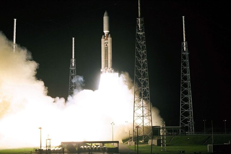 A tall, vertical rocket launches at night, its base surrounded by a massive, brightly glowing cloud of white smoke and intense yellow exhaust flames. The rocket is flanked by four tall, metal lattice lightning towers standing vertically against the solid black night sky. The brilliant white glare from the launch engines casts high-contrast light over the surrounding launchpad structures and ground.