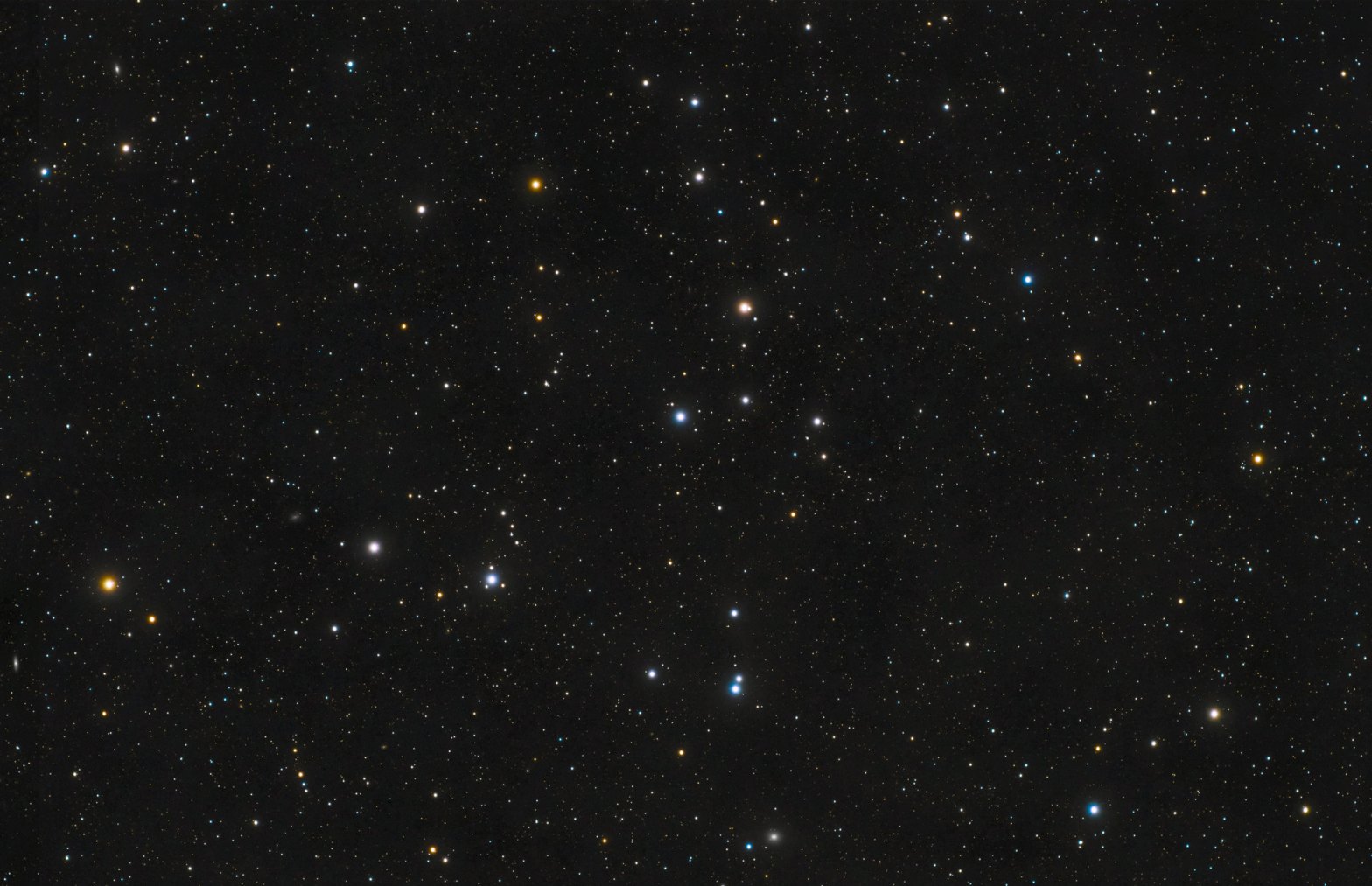 Telescopic photograph of the Coma Star Cluster (Melotte 111), showing a loose, scattered grouping of stars against a dense starfield. Stars display a range of colors including white, blue-white, and orange-gold, set against a black background.