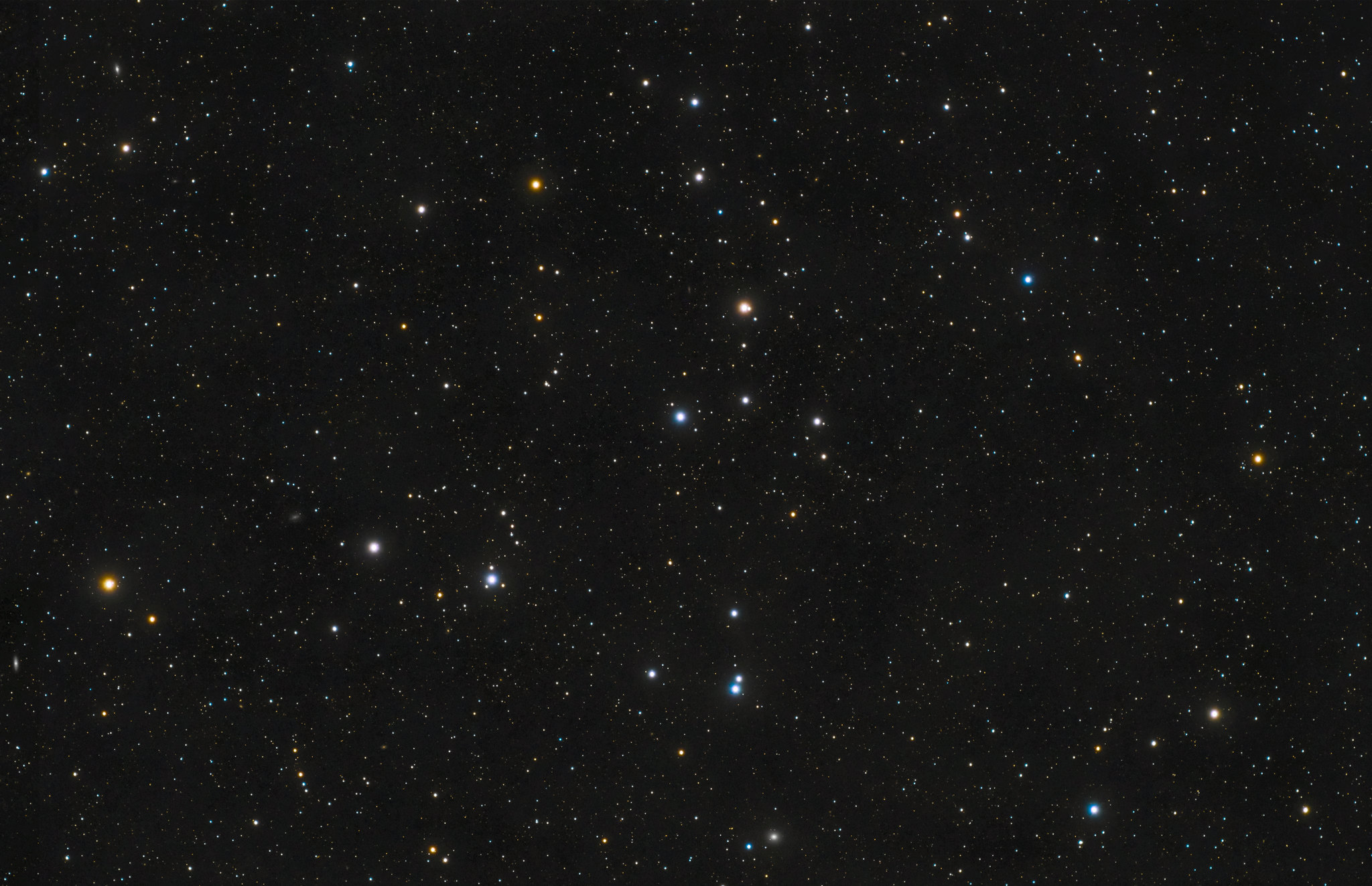 Telescopic photograph of the Coma Star Cluster (Melotte 111), showing a loose, scattered grouping of stars against a dense starfield. Stars display a range of colors including white, blue-white, and orange-gold, set against a black background.