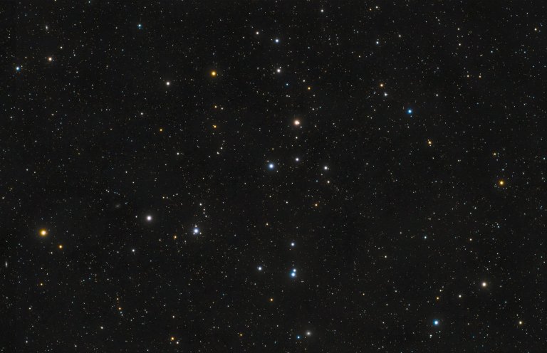 Telescopic photograph of the Coma Star Cluster (Melotte 111), showing a loose, scattered grouping of stars against a dense starfield. Stars display a range of colors including white, blue-white, and orange-gold, set against a black background.