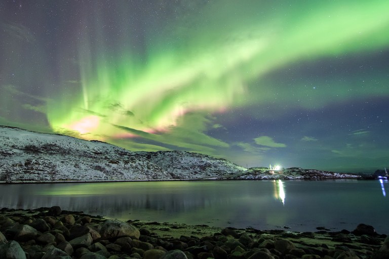A vivid aurora borealis fills the night sky with sweeping green and pink curtains of light above a snow-covered coastal hillside in the Arctic. The calm water of a fjord reflects the glow, while rounded rocks line the shoreline in the foreground and distant lights illuminate a small structure on the far shore.