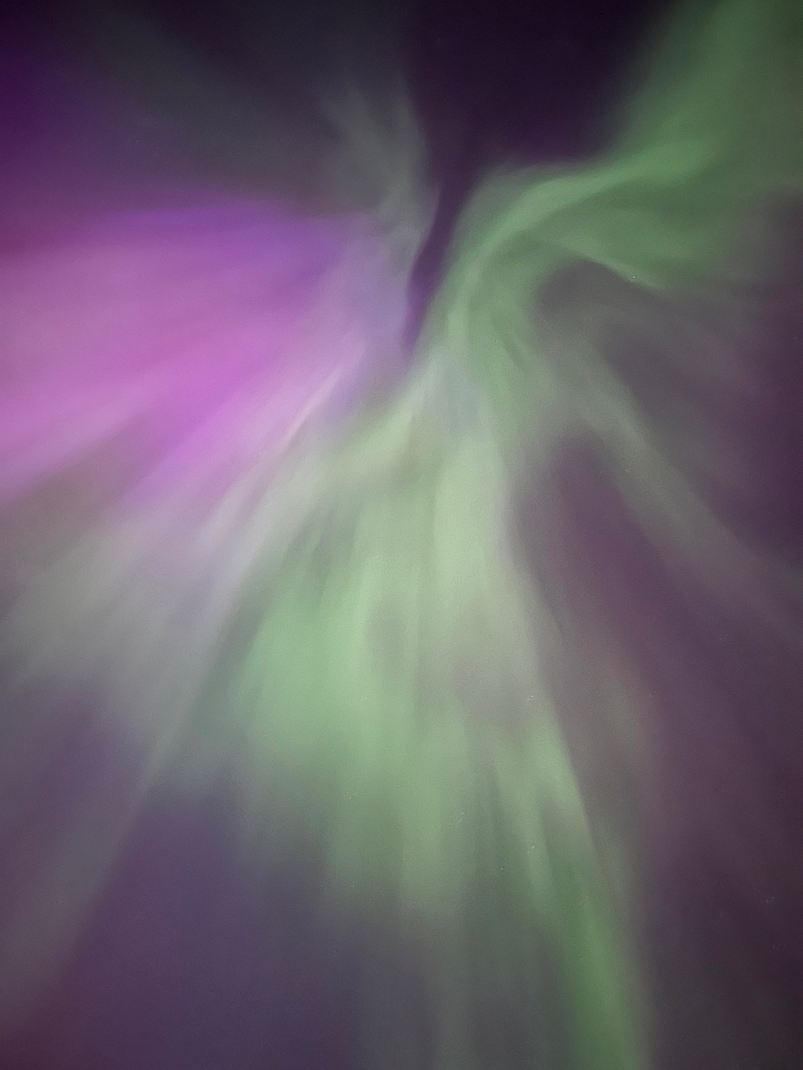 A photograph of an aurora borealis, with swirling curtains of pale green and violet light converging toward a central point overhead, filling the entire frame against a dark sky.