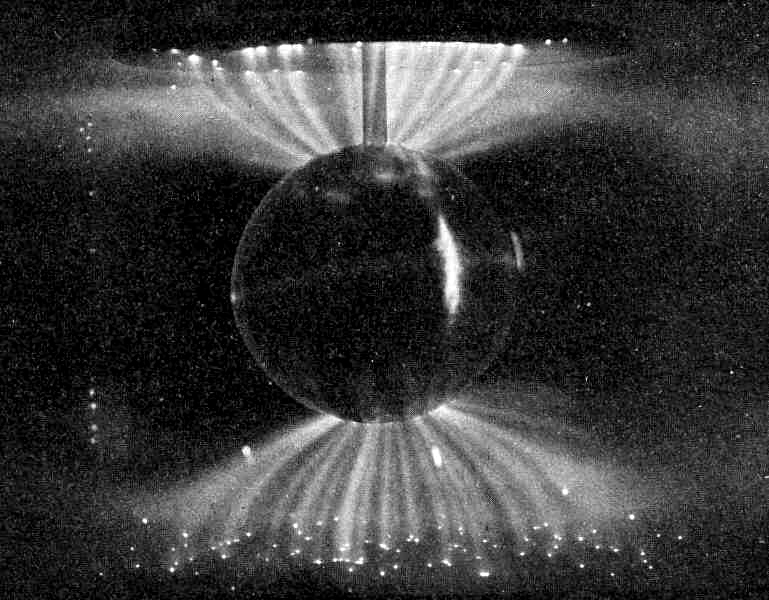 A black-and-white scientific photograph showing a dark sphere at center, meant to represent Earth, surrounded by symmetrical rays of electrical current streaming outward from both poles to demonstrate how aurorae are formed.