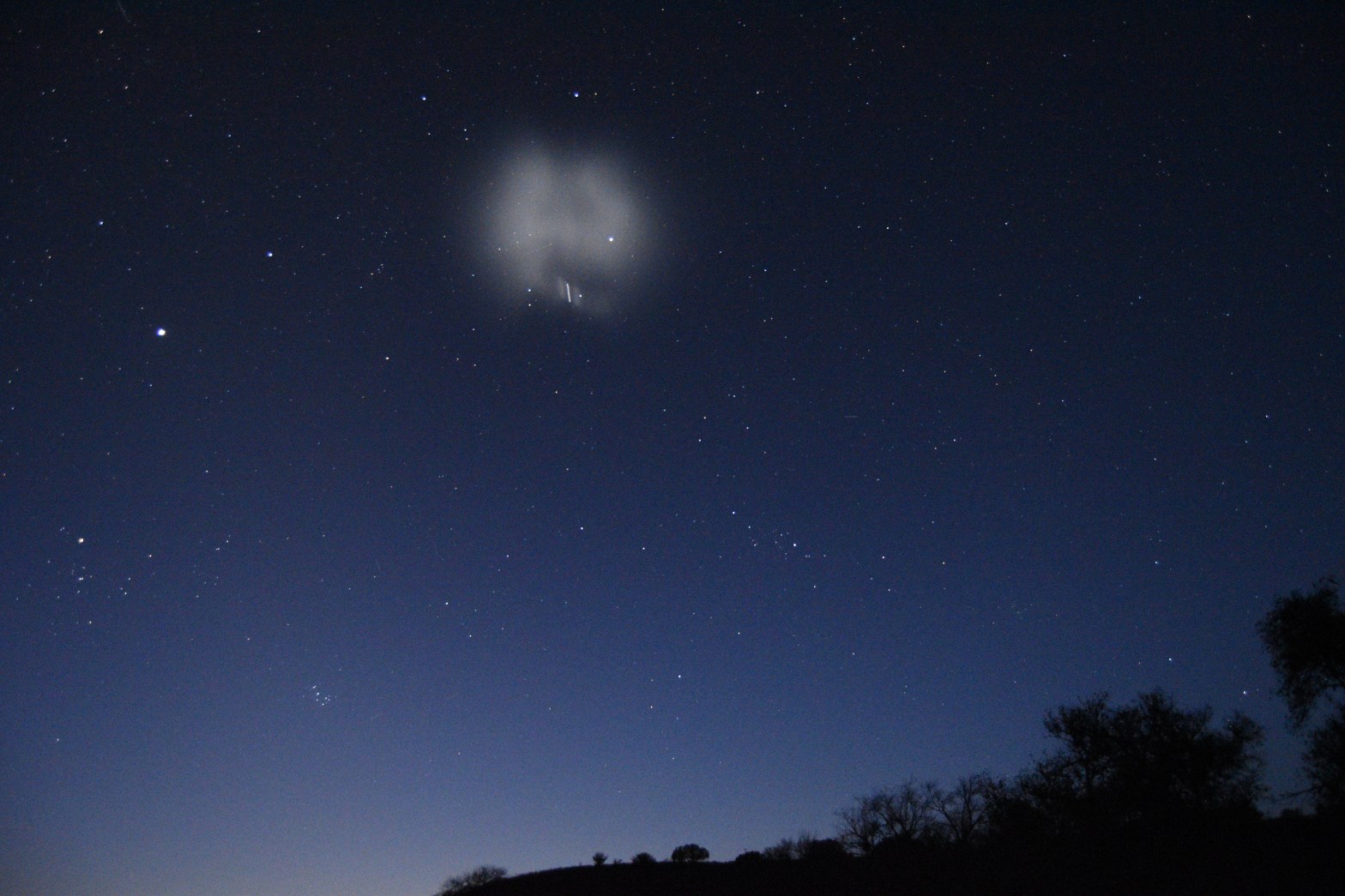 A wide-angle night sky photograph showing a glowing, diffuse white cloud created by a rocket fuel dump, billowing against a deep blue starry sky. A bright streak of light is visible within the expanding plume. Silhouetted trees and a hillside are visible along the bottom, and the sky fades to a lighter blue near the horizon.