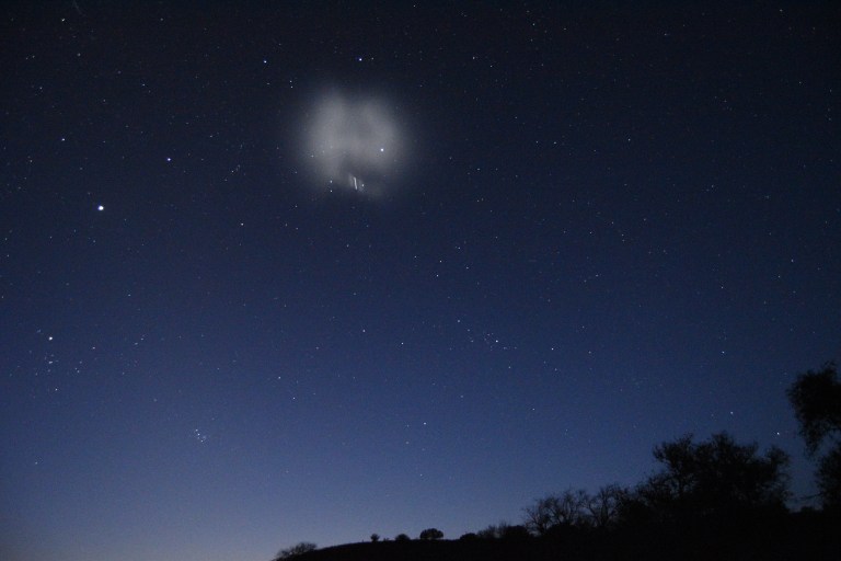 A wide-angle night sky photograph showing a glowing, diffuse white cloud created by a rocket fuel dump, billowing against a deep blue starry sky. A bright streak of light is visible within the expanding plume. Silhouetted trees and a hillside are visible along the bottom, and the sky fades to a lighter blue near the horizon.