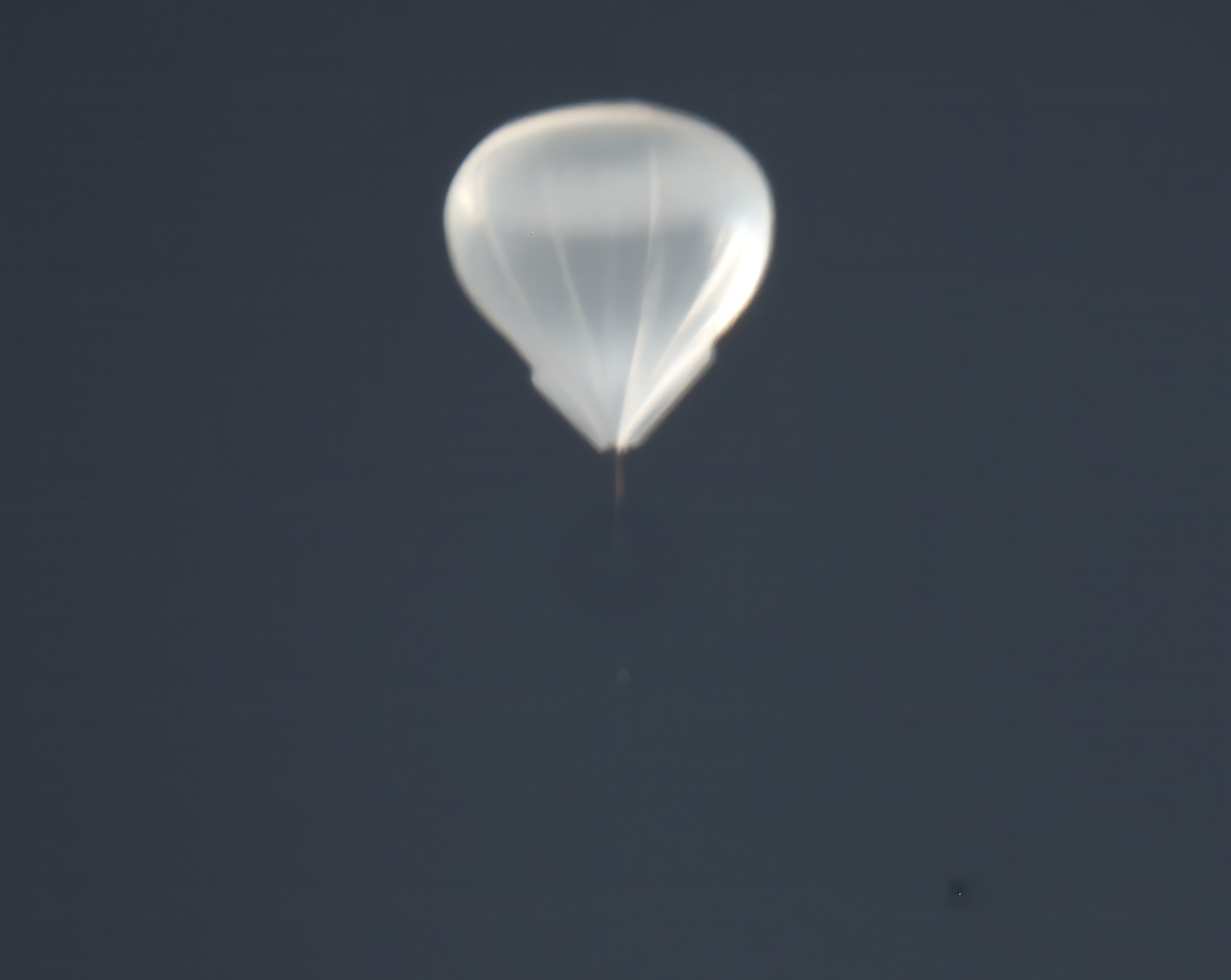 A close-up of a high-altitude balloon floating against a dark gray sky. The balloon's translucent white envelope is partially inflated into a rounded teardrop shape, with visible panel seams and a thin tether or payload line dangling below it.