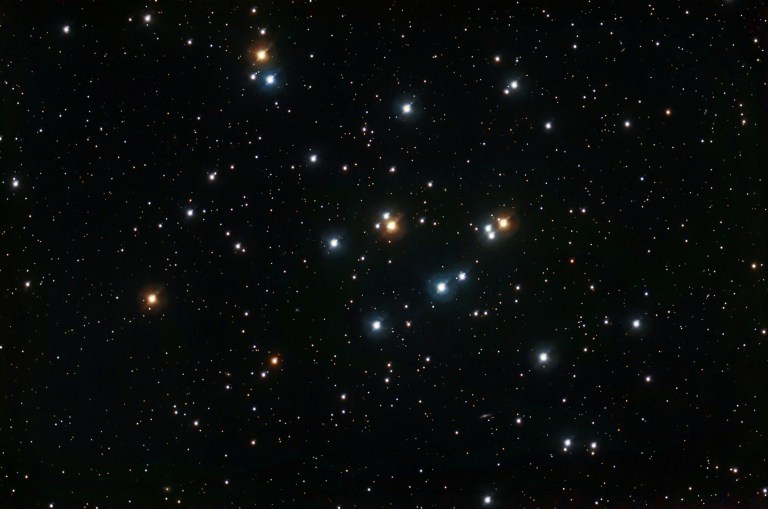 A wide-field view of M44, the Beehive Cluster, showing a loose, sprawling grouping of bright, individually resolved stars scattered across the center of the frame. The cluster's most prominent members are large, brilliant blue-white stars, contrasted by several warm orange and golden stars interspersed among them. The cluster has no tight central concentration, spreading openly across the field with a rich background of much fainter stars visible throughout the deep black sky.