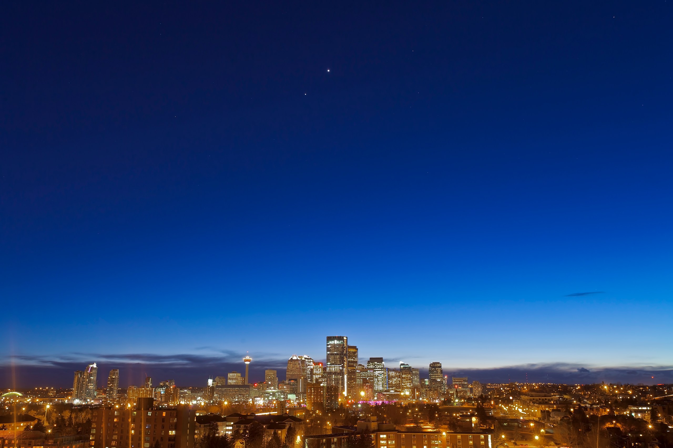 Two bright planets — Venus and Jupiter — shine as close points of light in a deep blue twilight sky above an illuminated city skyline at dusk. The city's skyscrapers, tower, and sprawling lights glow against a gradient sky that fades from deep blue above to lighter blue near the horizon, with low clouds hugging the rooftops.