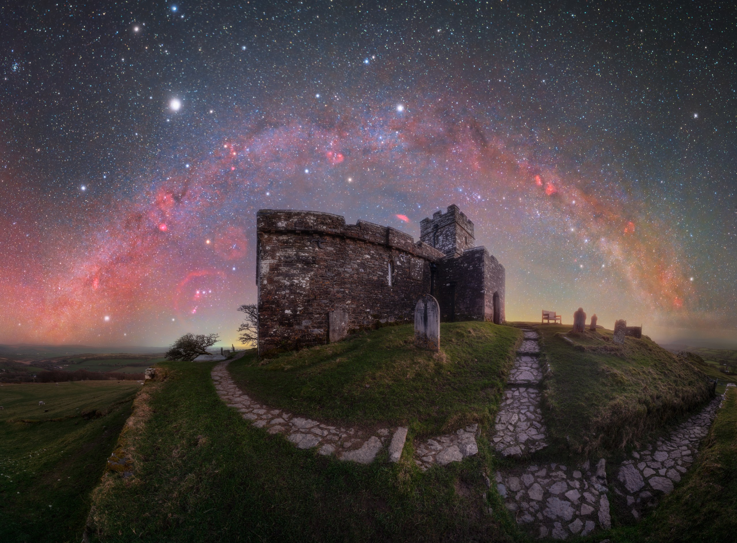 A dark, jagged hilltop topped by a small church stands against a glowing night sky filled with stars. The Milky Way arches high overhead as a bright, dense band of light, with streaks and clumps of dark dust and red gas fading into a softer, mottled glow. The horizon is lit by a faint warm haze, separating the dark foreground from the richly textured starfield above.