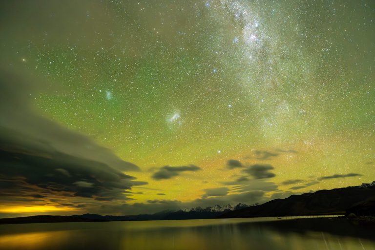 Bands of soft green aurorae stretch above a jagged, silhouetted mountain ridge under a star-filled sky, reflecting off a body of water in the foreground. The Milky Way forms a bright, hazy band stretching from the right of the frame to the top. Beside it at left lie a few compact, fuzzy light patches — the Small and Large Magellanic Clouds.