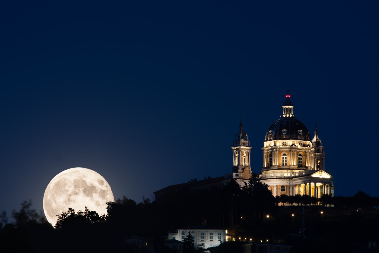 A large Full Moon rises behind trees at left, glowing bright white against a deep blue night sky. To the right, the illuminated Baroque dome and bell tower of the Basilica of Superga in Turin, Italy are lit in warm golden light, creating a dramatic contrast with the moonlit sky.