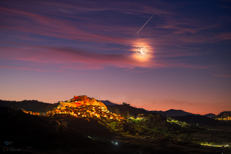 A single bright meteor streaks diagonally across a dark, star-filled sky above a rugged hilltop town. The lights and stone buildings of the town cluster around a rocky ridge, with a soft, pink glow near the horizon. A crescent Moon is enveloped by scattered clouds, generating a bright, wispy halo.