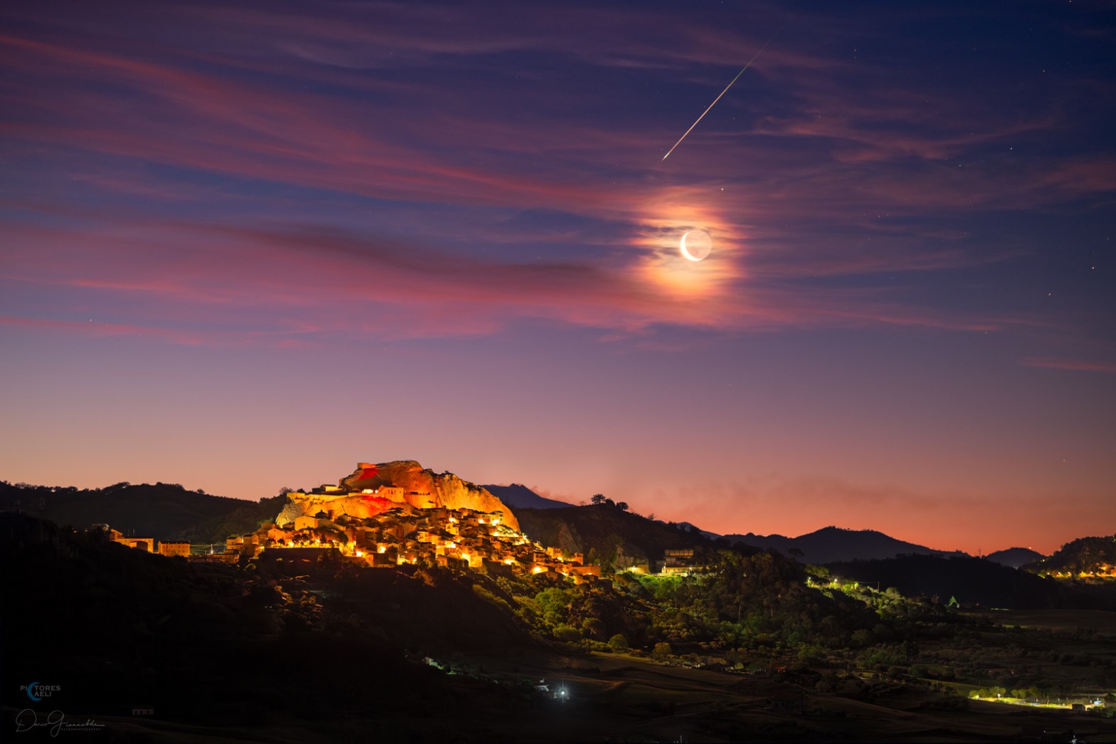 A single bright meteor streaks diagonally across a dark, star-filled sky above a rugged hilltop town. The lights and stone buildings of the town cluster around a rocky ridge, with a soft, pink glow near the horizon. A crescent Moon is enveloped by scattered clouds, generating a bright, wispy halo.