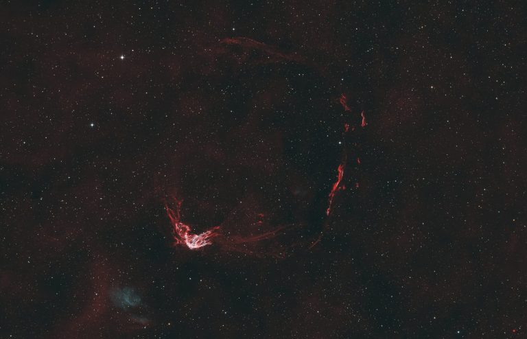 A wide, tangled ring of thin, reddish filaments wraps around a darker central region, forming an uneven circle against a black sky. The ring is mottled and wispy, with brighter knots and fainter strands that fade into the background. Myriad small stars of varying brightness are scattered across the frame and in front of the ring.