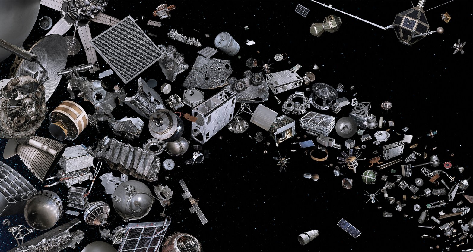 A dense cluster of silver and gray spacecraft components, rocket parts, and satellite fragments floats against a black starfield, packed tightly on the left side of the frame and thinning into scattered individual pieces toward the right. The objects range from large cylindrical rocket stages and solar panel arrays to small bolts and twisted metal fragments.