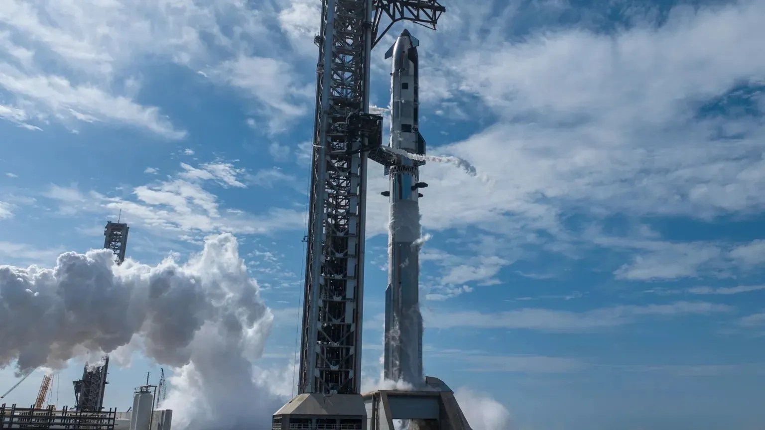 SpaceX's Starship rocket stands fully stacked on its launch mount at Boca Chica, Texas, with the towering mechazilla catch arm structure rising alongside it on the left. Thick white clouds of venting propellant or water vapor billow around the base and drift to the left, while a blue sky with scattered white clouds fills the background. A second launch structure is visible in the far left of the frame.