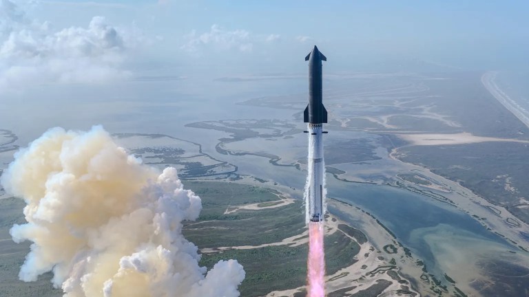 SpaceX's Starship rocket climbs away from its launch site shortly after liftoff, its white booster stage trailing a bright pink and white flame plume downward toward a coastal wetlands landscape below. A large billowing cloud of yellow-white exhaust smoke rises to the left of the vehicle, while winding waterways, sandy shores, and patches of green vegetation stretch across the terrain beneath a pale blue sky.