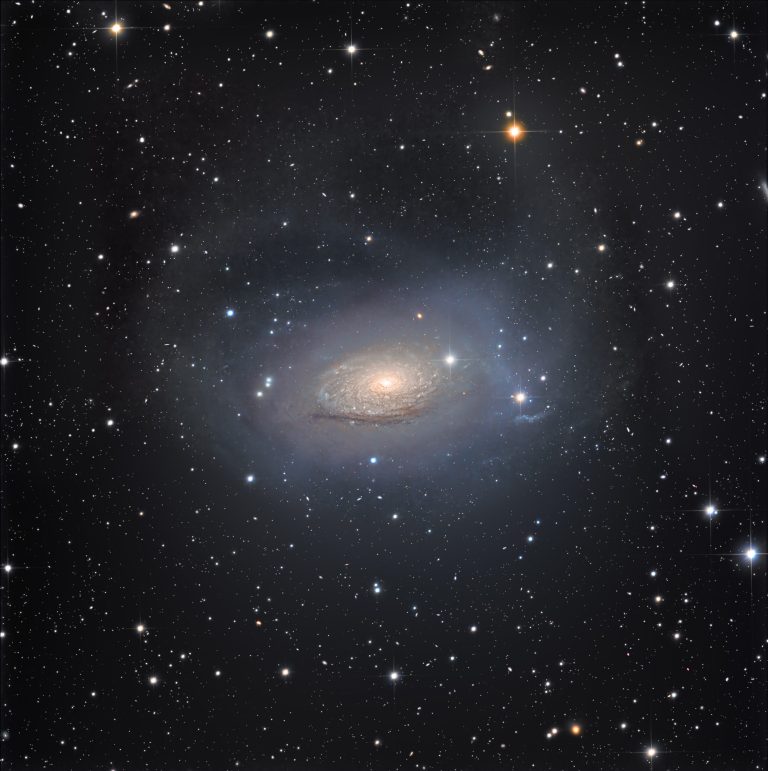 A deep image of the Sunflower Galaxy set against a dark sky, with a bright, compact core wrapped in soft, uneven spirals of light. Very faint, misty arcs and streaks of starlight extend far beyond the main disk, forming a large, irregular halo around the galaxy. The background is sprinkled with small, sharp stars in white, blue, and amber, along with a few tiny, hazy distant galaxies.