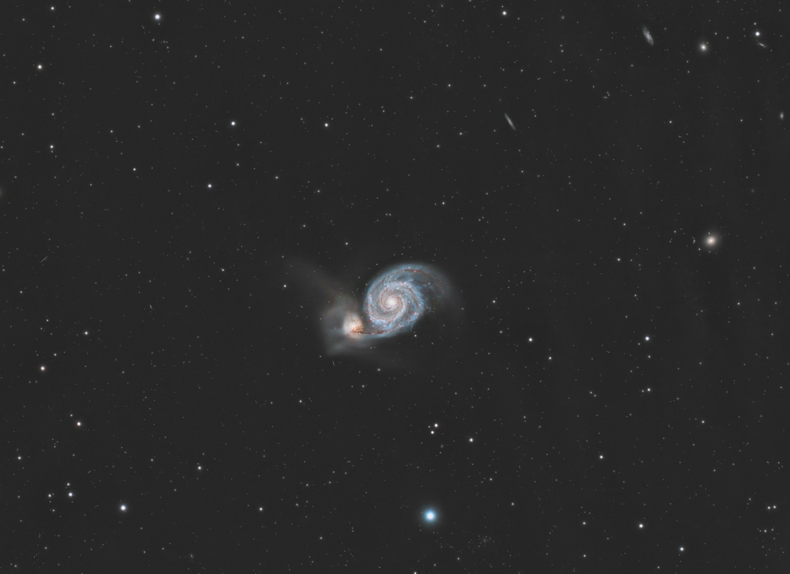 A face-on spiral galaxy sits near the center of the frame, its bright, compact core surrounded by two broad, curving arms mottled with pale blue and pinkish patches. A smaller, hazier companion galaxy clings to the outer edge of one arm, slightly offset and to the side, where their halos appear to blend. The scene is set against a dark background sprinkled with faint foreground stars and a few tiny, distant galaxies.