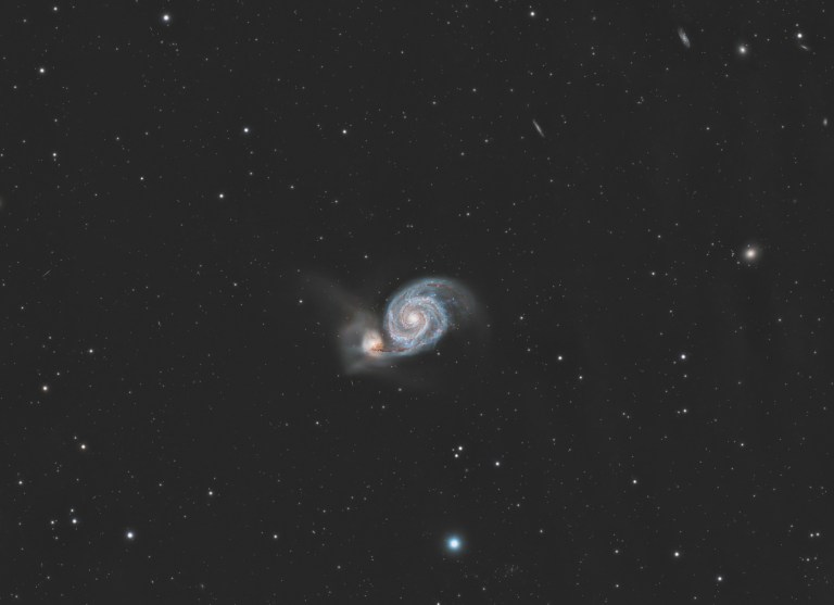A face-on spiral galaxy sits near the center of the frame, its bright, compact core surrounded by two broad, curving arms mottled with pale blue and pinkish patches. A smaller, hazier companion galaxy clings to the outer edge of one arm, slightly offset and to the side, where their halos appear to blend. The scene is set against a dark background sprinkled with faint foreground stars and a few tiny, distant galaxies.