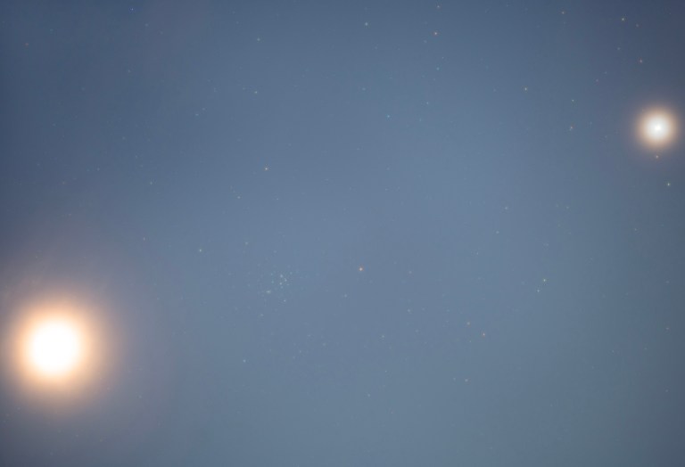The warm, overexposed Moon glows in the lower left of a blue-gray twilight sky, while a smaller, intensely bright Venus shines in the upper right. Between them, a loose grouping of faint stars — the Beehive Cluster — is just barely visible near the center of the frame against the hazy background.