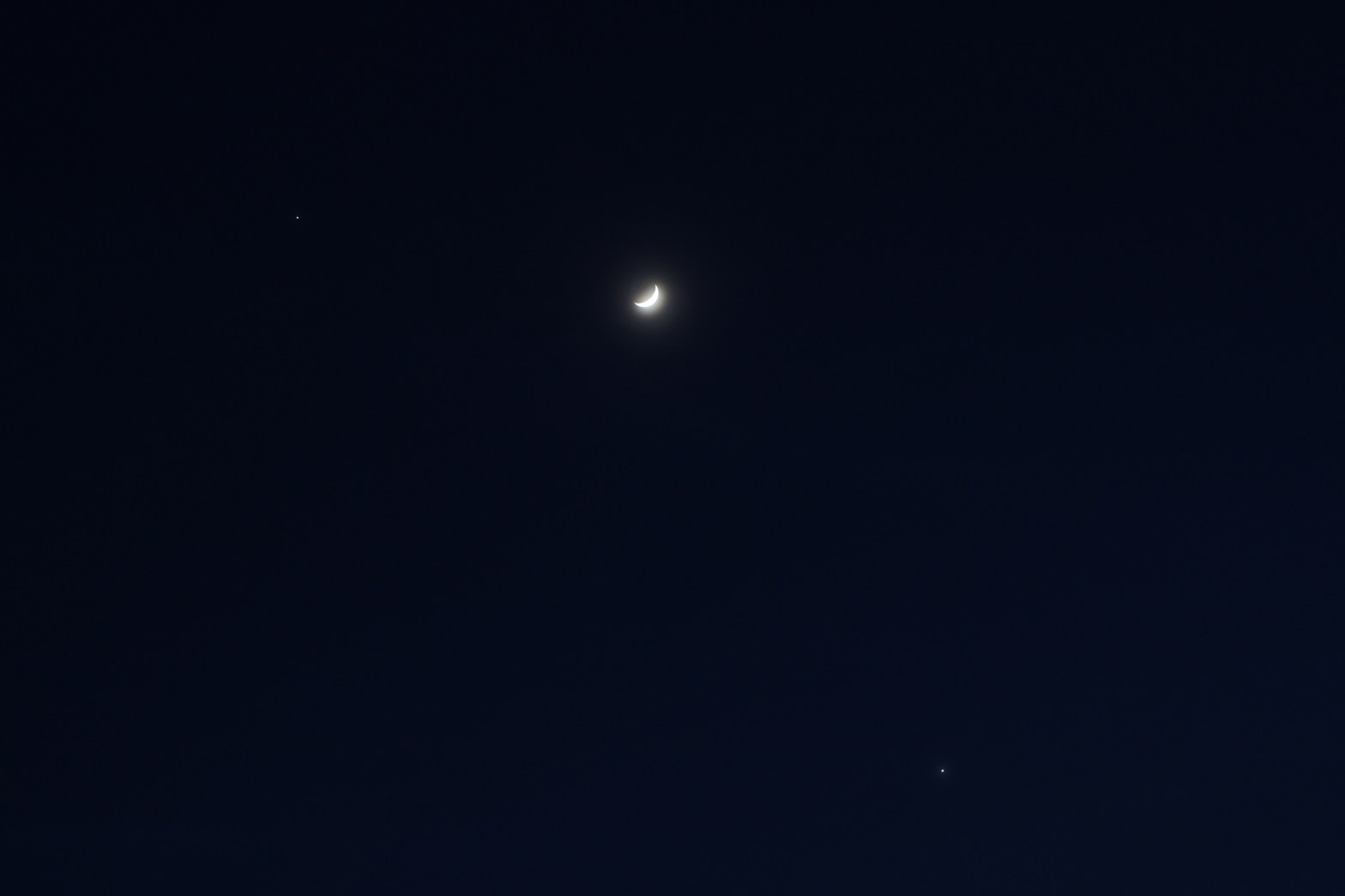 A crescent moon glowing brightly against a deep dark blue night sky, flanked by two planets, Jupiter and Venus, visible as small bright points — one to the upper left and one to the lower right.