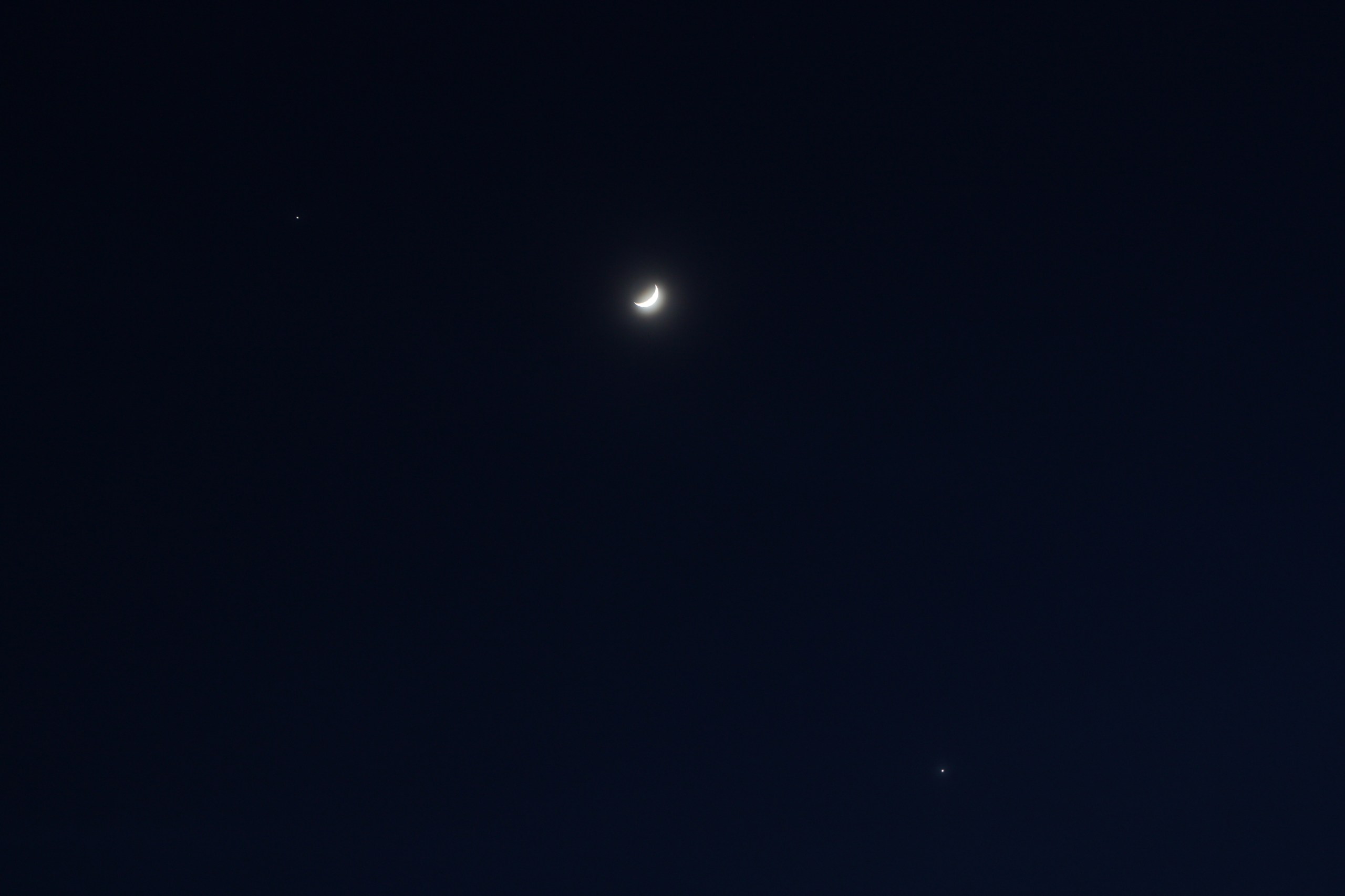 A crescent moon glowing brightly against a deep dark blue night sky, flanked by two planets, Jupiter and Venus, visible as small bright points — one to the upper left and one to the lower right.