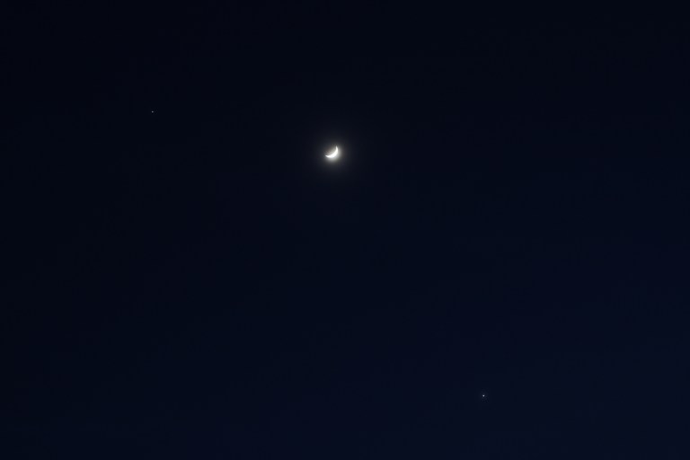 A crescent moon glowing brightly against a deep dark blue night sky, flanked by two planets, Jupiter and Venus, visible as small bright points — one to the upper left and one to the lower right.