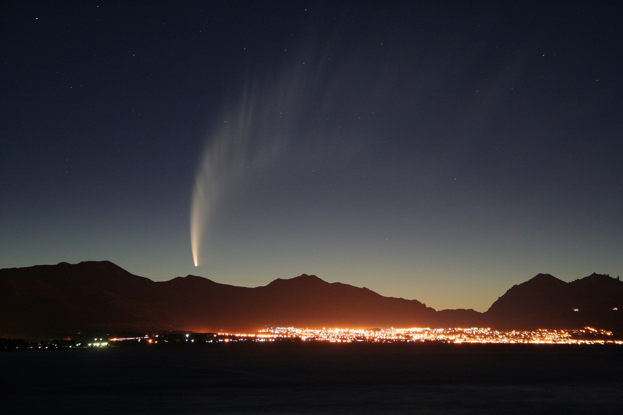 A bright comet with a long tail over mountains and city lights
