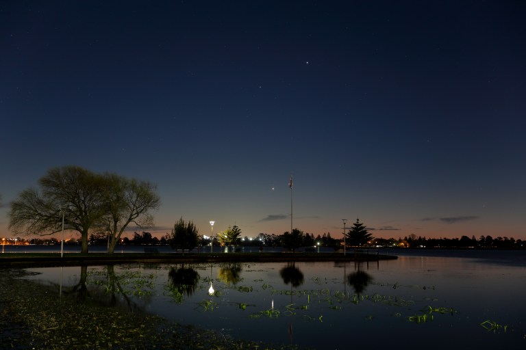 A twilight photograph taken from the shore of a calm lake, looking toward a park with trees, streetlights, and a tall flagpole silhouetted against a deep blue and orange dusk sky. Three planets — Venus, Mercury, and Jupiter — are visible as bright points of light in the sky, with two clustered close together near the horizon and one higher up. Their reflections shimmer faintly in the still water below, which is dotted with aquatic plants.