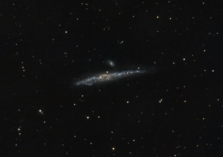 A telescopic photograph of the Whale Galaxy (NGC 4631), an edge-on spiral galaxy appearing as a long, thin streak of blue and white light with a bright, mottled core, set against a black sky filled with foreground stars. A smaller companion galaxy is visible just above the main galaxy near the center of the image.