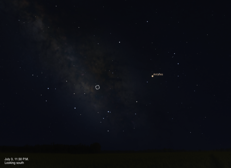 A simulated night sky looking south on July 3, 2026 at 11:30 P.M. A small circular target indicator marks an unnamed object embedded in the faint glow of the Milky Way near the center of the frame. The bright orange-red star Antares is visible to the right, and the dense star clouds of the Milky Way stretch across the left side of the image.