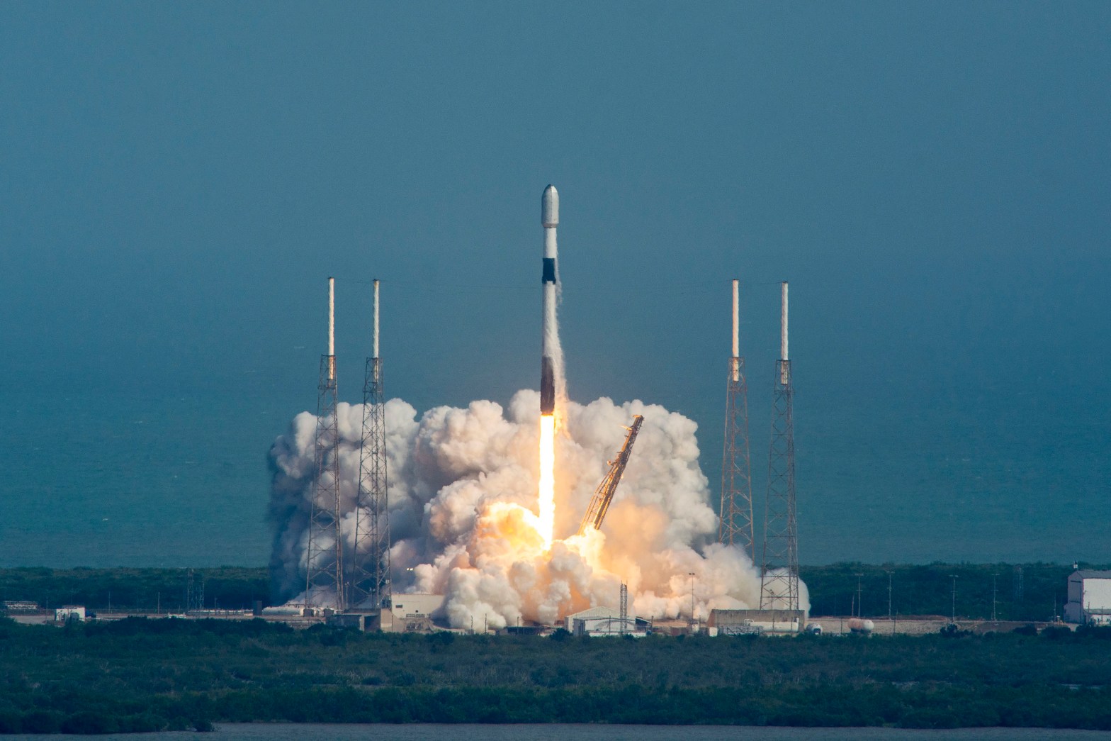 A SpaceX Falcon 9 rocket rises from its launch pad at Cape Canaveral Space Force Station, surrounded by a billowing cloud of white exhaust and a bright orange flame at the base. The rocket climbs vertically against a hazy blue-gray sky, flanked by four lightning rod towers. Launch infrastructure, including a yellow crane arm and concrete support structures, is visible at the bottom of the frame.