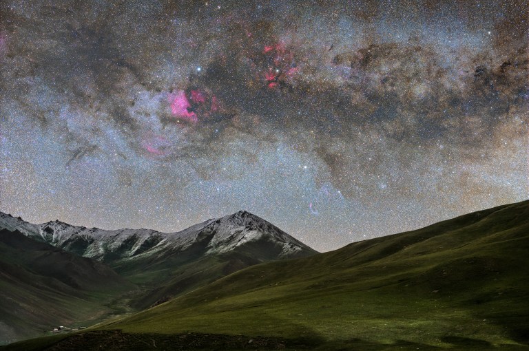 The Milky Way stretches in a broad arc across a dark, star-filled sky above a mountain range with snow-dusted peaks and green alpine valleys. Bright pink and magenta clouds of glowing gas are visible in the upper left portion of the sky, with patches of dark dust threading through the dense band of stars. A few small structures — including a tiny settlement — are visible at the base of the mountains.